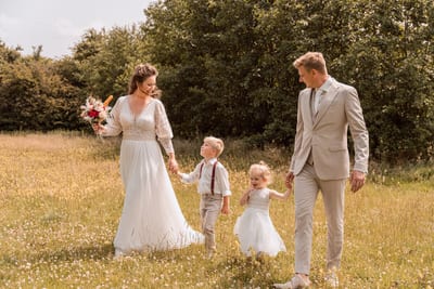 Bert en Verena wandelen hand in hand met twee kinderen in feestkleding door een zomerse bloemenweide op hun trouwdag