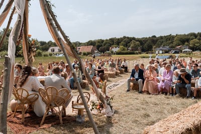 Bert en Verena zitten tijdens hun buitenceremonie onder een houten tipi, terwijl gasten op strobalen in het veld toekijken