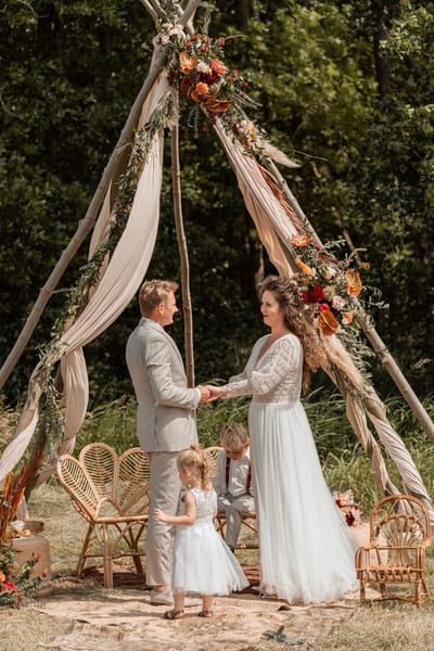 Bert en Verena houden elkaars handen vast onder een met doeken en bloemen versierde tipi tijdens hun buitenceremonie
