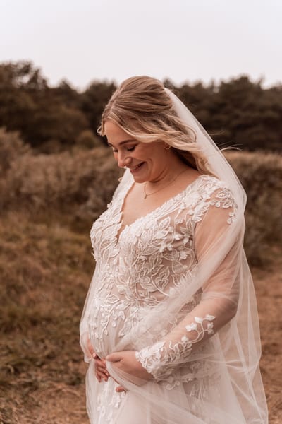 Bruid Femke kijkt glimlachend omlaag met haar handen om haar buik, haar sluier waait over de kanten jurk in het open veld