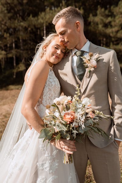 Esmee leunt met gesloten ogen tegen Danny's schouder, het veldboeket met droogbloemen in haar handen, in warm zonlicht