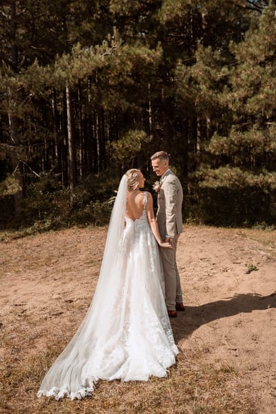 Danny en Esmee staan hand in hand aan de rand van een naaldbos, de kanten sleep uitgespreid over het zand