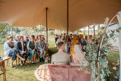 Danny en Esmee zitten tijdens de ceremonie samen op een roze bankje onder een stretchtent, met alle gasten tegenover hen