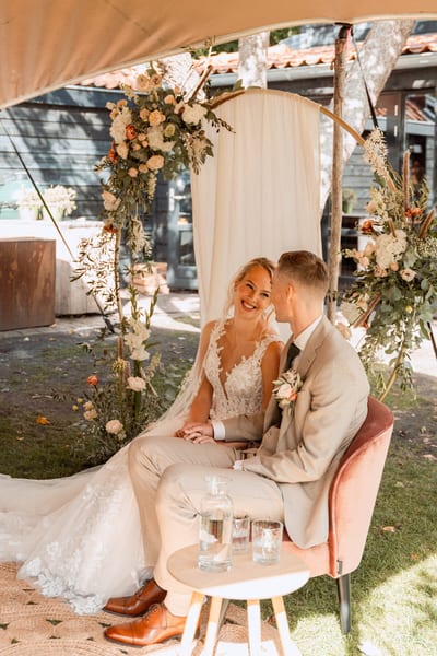 Danny en Esmee zitten lachend naast elkaar op een roze fluwelen bank voor een bloemenboog, een karaf water op het tafeltje voor hen