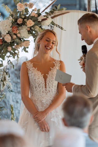 Esmee lacht stralend terwijl Danny zijn geloften voorleest bij de bloemenboog tijdens de ceremonie