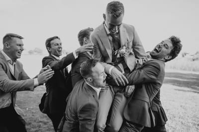 Danny wordt door zijn lachende vrienden opgetild tijdens een uitgelaten moment op de trouwdag, in zwart-wit