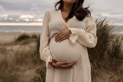 Zwangere vrouw omvat haar buik met beide handen tussen het helmgras in de duinen, de zee op de achtergrond