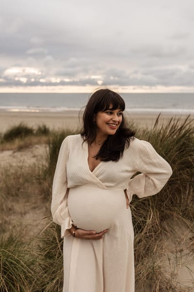 Zwangere vrouw in crèmekleurige jurk houdt haar buik vast tussen het helmgras, met de zee onder een dramatische wolkenlucht