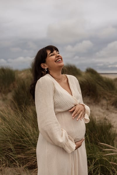 Zwangere vrouw in crèmekleurige jurk lacht met haar hoofd in de nek tussen het helmgras in de duinen