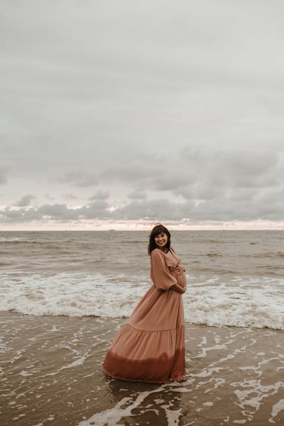 Zwangere vrouw staat in haar terracotta jurk met de handen om haar buik in de branding, onder een dramatische wolkenlucht