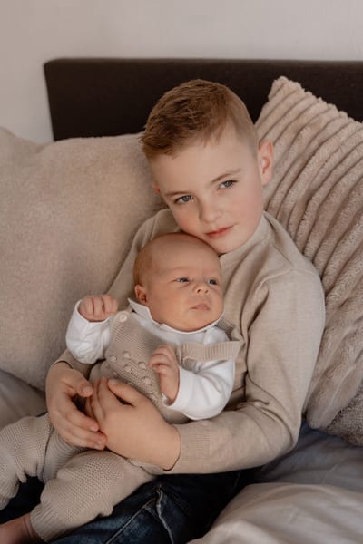 Grote broer houdt de pasgeboren baby trots op schoot tussen zachte kussens op bed