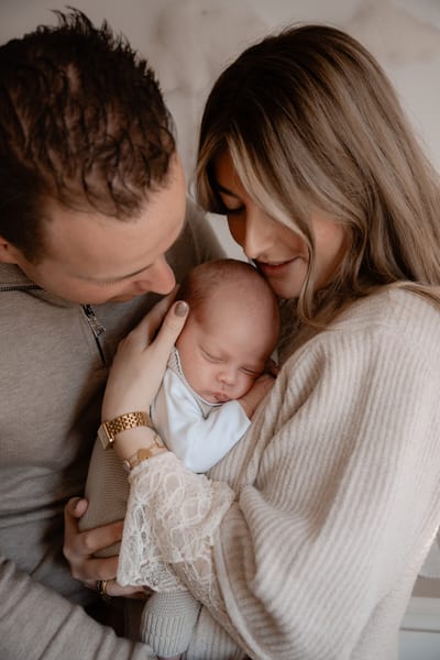 Ouders buigen hun hoofden naar elkaar toe over hun slapende newborn in de armen van de moeder, in zachte lichte tinten