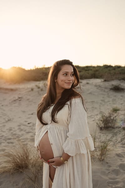 Aanstaande moeder toont haar blote buik in een crèmekleurige jurk tussen de duinen, in het gouden licht van de ondergaande zon