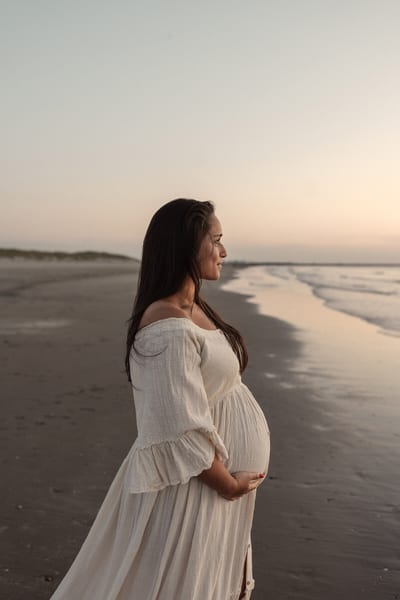 Zwangere vrouw kijkt bij zonsondergang uit over zee, haar handen rustend onder haar buik op het strand