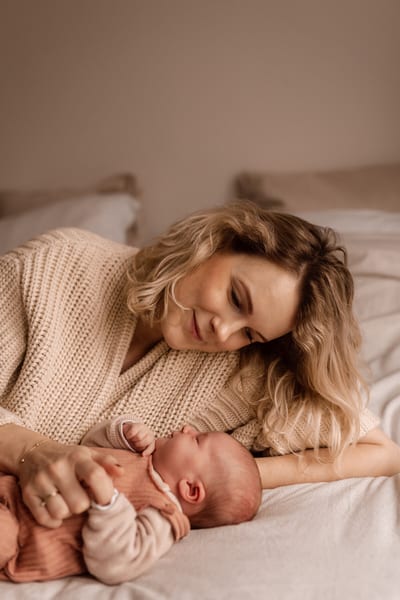 Moeder ligt op bed en kijkt vertederd naar haar pasgeboren baby, hun handen liggen in elkaar