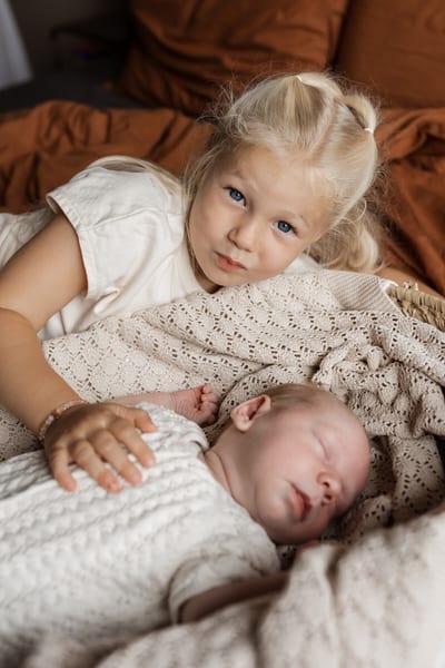 Grote zus kijkt in de camera terwijl ze haar hand beschermend op de slapende baby legt, tussen zachte gebreide dekens
