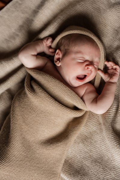Pasgeboren baby gaapt met beide armpjes omhoog, gewikkeld in een zachte gebreide deken