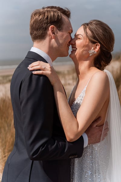 Mart en Loes staan lachend neus aan neus in de duinen, met het strand en de zee op de achtergrond