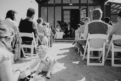 Terwijl Mart en Loes bij het strandhuis hun ceremonie beleven, speelt een kindje in het zand tussen de gastenstoelen