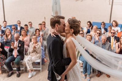 Mart en Loes kussen elkaar op het strand terwijl de sluier opwaait en de gasten zittend applaudisseren