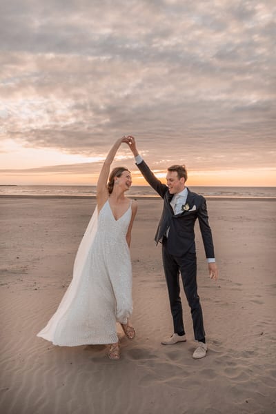 Bruidegom draait de bruid lachend onder zijn arm tijdens een dans op het strand bij zonsondergang