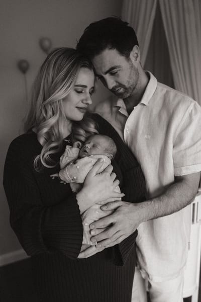 Ouders staan hoofd aan hoofd met hun pasgeboren baby in de armen, zwart-witportret bij zacht licht in de slaapkamer