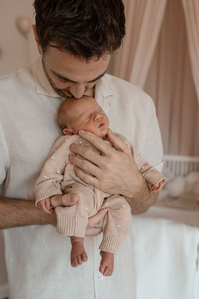Vader drukt een kus op het hoofdje van zijn pasgeboren baby, die in zijn handen slaapt in de zachtroze babykamer