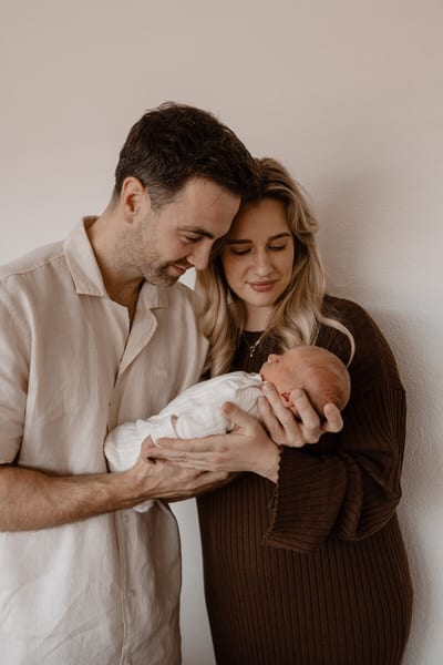 Ouders kijken samen liefdevol naar hun pasgeboren baby die in hun armen ligt, tegen een rustige lichte achtergrond
