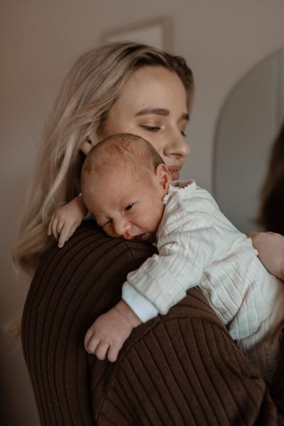 Pasgeboren baby ligt wakker op moeders schouder, weggedoken tegen haar bruine gebreide trui, in warm licht