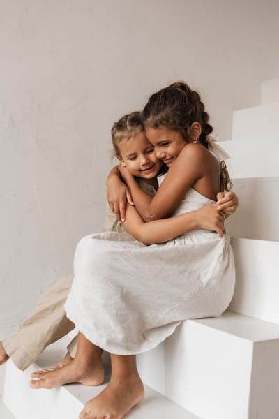 Twee meisjes omhelzen elkaar met gesloten ogen op een witte trap, blootsvoets in lichte zomerkleding tijdens de gezinsshoot