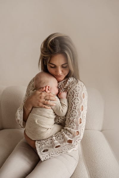 Moeder zit op een lichte bank en houdt haar slapende baby dicht tegen haar wang gedrukt