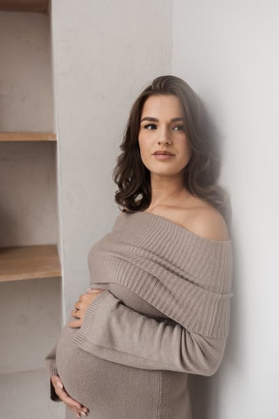 Zwangere vrouw in gebreide off-shoulderjurk leunt tegen een lichte wand met haar armen om haar buik