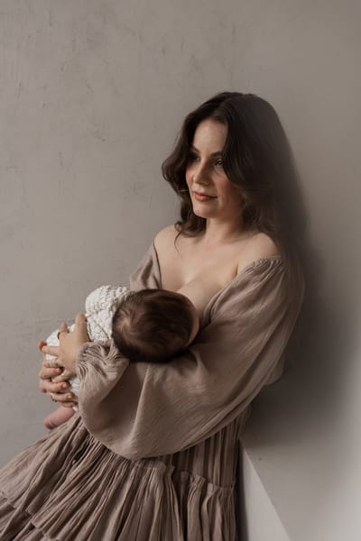Moeder in een zachte taupe jurk wiegt haar baby met gebreid mutsje in haar armen tegen een grijs gestucte wand