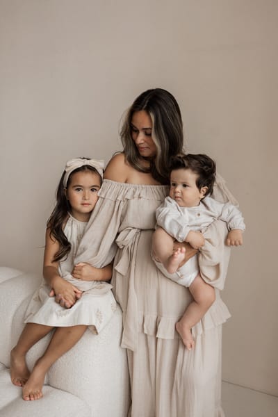 Moeder in beige jurk houdt haar baby op de arm terwijl haar dochter naast haar op de bank zit