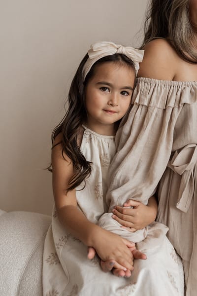Meisje met strik in het haar leunt tegen haar moeder aan en houdt haar arm vast, in zachte beige tinten