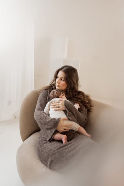 Moeder wiegt haar baby tegen haar borst in een ronde fauteuil, in zacht licht bij het raam
