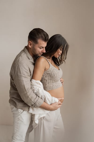 Partner omarmt de zwangere vrouw van achteren, samen leggen ze de handen op haar blote buik, in beige studiotinten