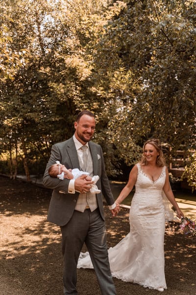 Rob draagt een baby in het wit op de arm terwijl hij en Tamara lachend door een zonnige tuin lopen op hun trouwdag