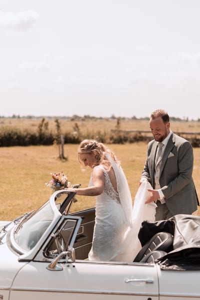 Tamara stapt met haar boeket in een klassieke witte cabriolet terwijl Rob haar jurk vasthoudt