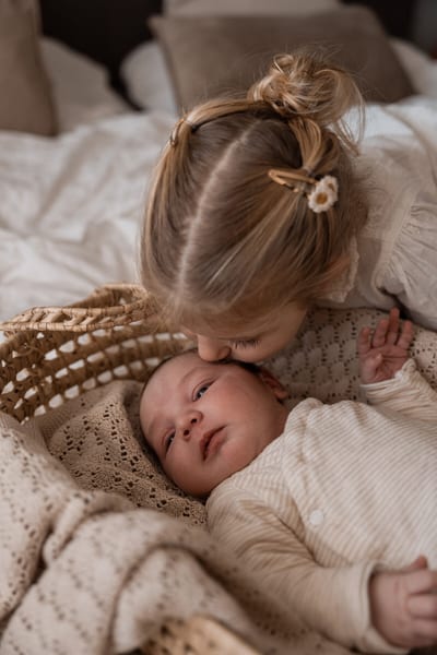 Grote zus geeft een kus op het hoofd van de newborn die in een rieten wiegje tussen zachte dekens ligt