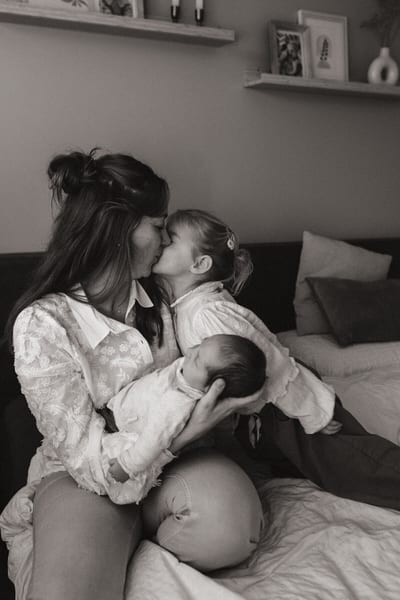 Grote zus kust haar moeder die de pasgeboren baby op schoot houdt op het bed, intiem gezinsmoment in zwart-wit