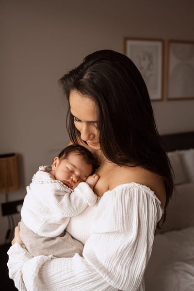 Moeder buigt zich liefdevol over haar slapende newborn, die tegen haar borst ligt gewikkeld in een warme slaapkamer