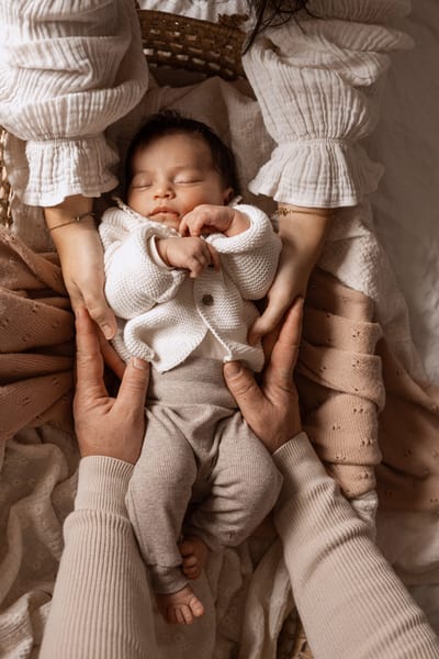 Slapende newborn in een wit gebreid vestje, omringd door de handen van beide ouders in een rieten mandje