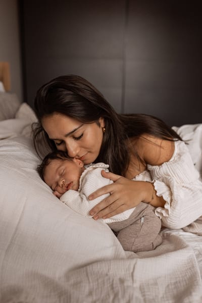 Moeder buigt zich op bed liefdevol over haar slapende baby en houdt het kindje dicht tegen zich aan