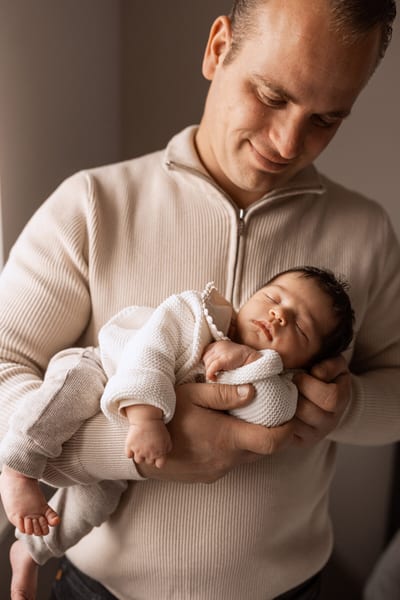 Vader kijkt glimlachend neer op zijn slapende pasgeboren baby met donker haar, in zijn armen bij zacht raamlicht