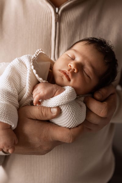 Pasgeboren baby slaapt in een gebreid pakje, veilig omsloten door de handen van een ouder in warm licht