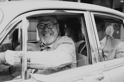 Lachende chauffeur met vlinderdas rijdt Thijs en Lois in een klassieke trouwauto, vastgelegd in zwart-wit