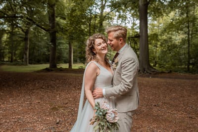 Thijs en Lois staan lachend wang aan wang in het bos, een boeket met grote protea in haar hand