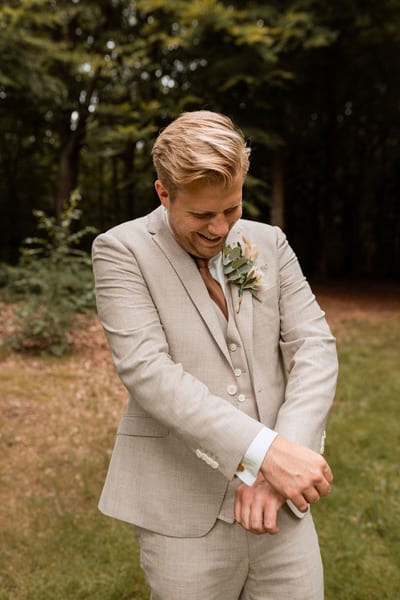 Thijs schikt lachend de manchet van zijn beige driedelige trouwpak aan de rand van een groen bos
