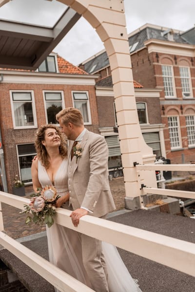 Thijs en Lois staan dicht bij elkaar op een witte ophaalbrug, Lois kijkt lachend omhoog met haar boeket in de hand